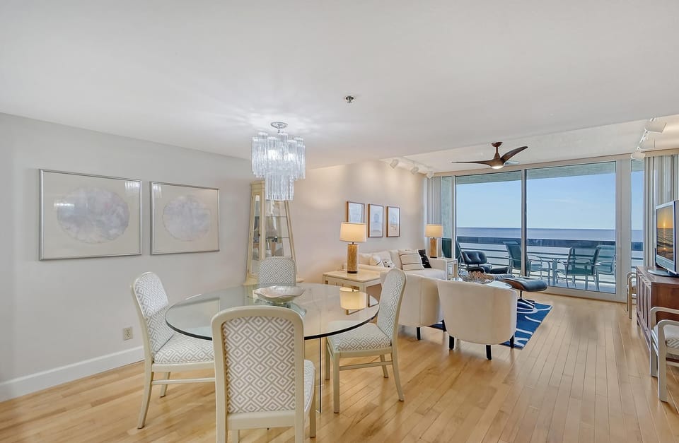 Round glass dining table surrounded by patterned white chairs, topped with a tiered glass chandelier.