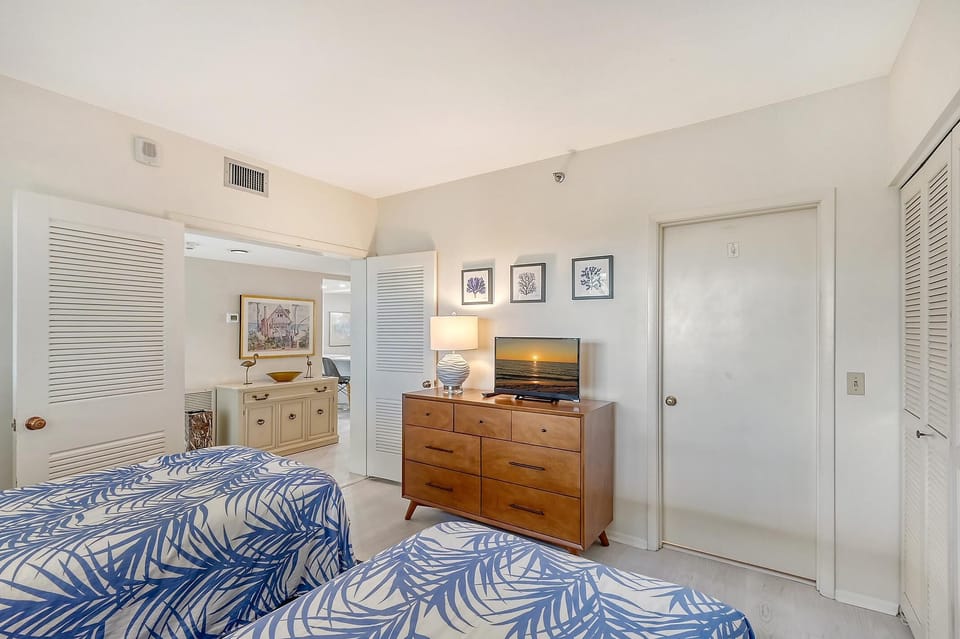 Twin beds with blue leaf-patterned bedding face a wood dresser topped with a flat-screen TV and lamp.