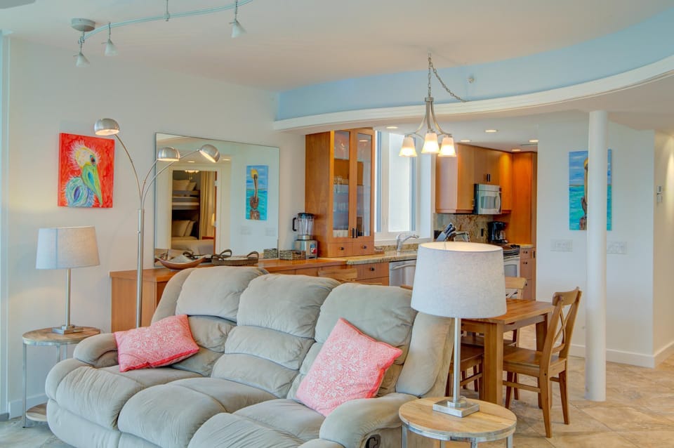 Open-concept living and dining area with a plush gray sofa, wooden table, and kitchen featuring warm wood cabinets.