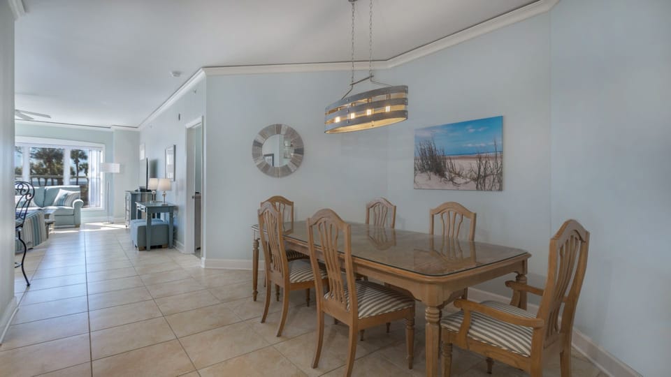 The glass-top table seats six beneath a modern coastal light fixture, with soft neutral tones and crown molding adding a polished finish.