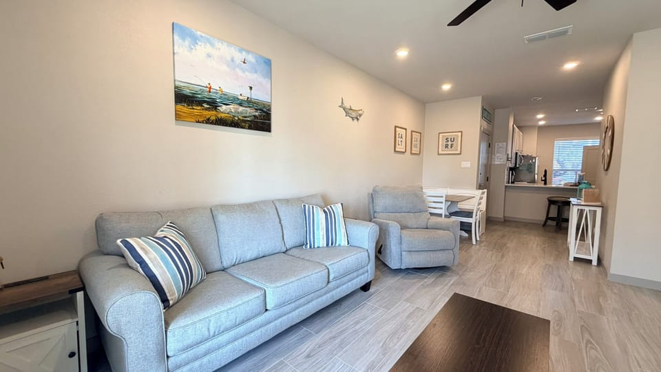 Your spacious living room invites relaxation with its comfortable grey sofa, coastal artwork, and open-concept flow into the kitchen.