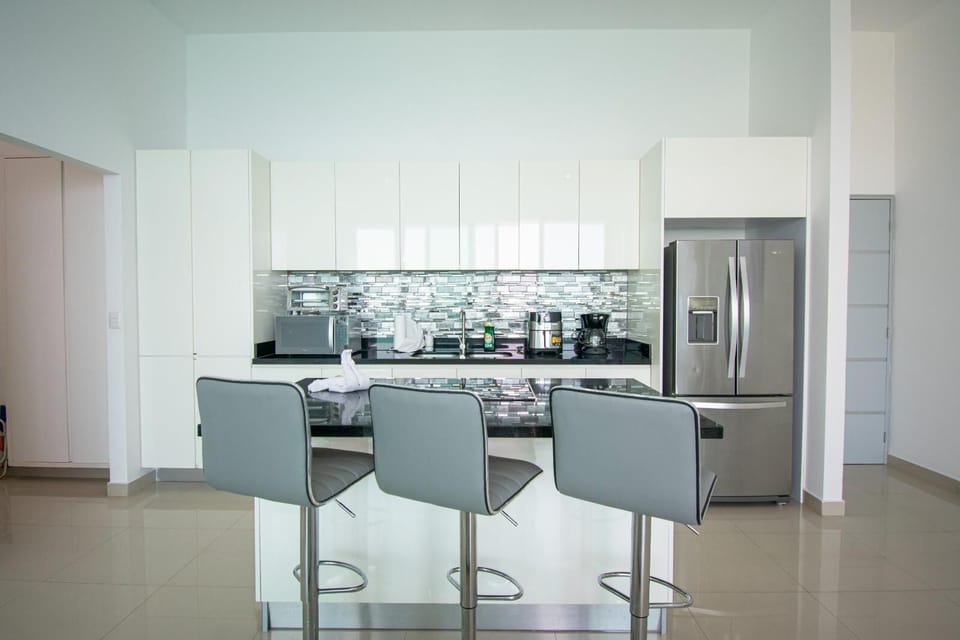 Glossy white cabinets and a reflective silver tile backsplash contrast with the black granite island topped by three gray barstools.