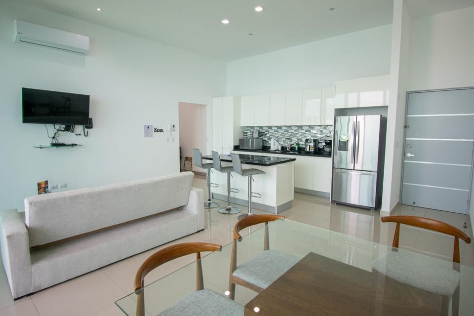 Glass dining table with wooden chairs overlooks a white and black kitchen featuring a tiled backsplash and stainless steel fridge.