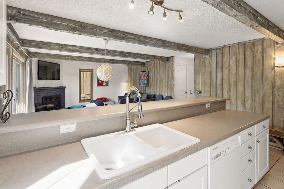 A kitchen with a white double sink and light gray countertops opens to a living area with a fireplace and multiple seating options. The ceiling has exposed wooden beams and a modern light fixture.
