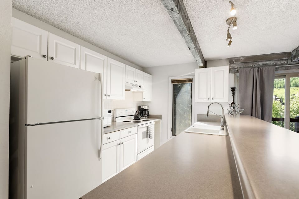 A modern kitchen with white cabinets, a refrigerator, stove, microwave, sink, and track lighting. A window overlooks greenery outside. Counters are clean and uncluttered.