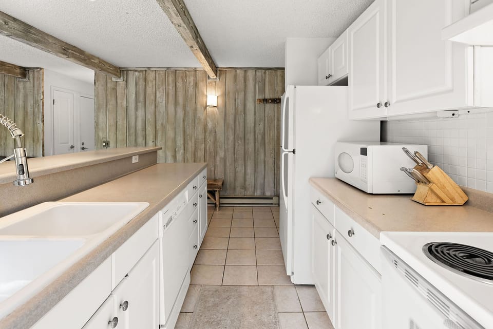 A white modern kitchen features wooden beams, white cabinetry, a refrigerator, a microwave, a sink, a dishwasher, and a countertop with a knife block. Walls are lined with vertical wooden slats.