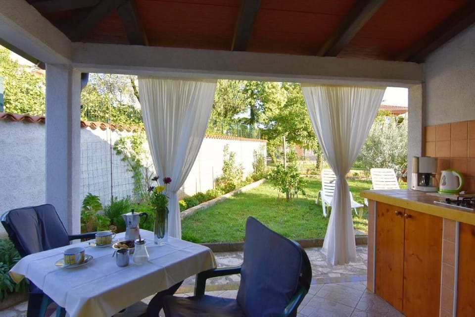 The covered terrace has an outdoor kitchen, table with 2 chairs, awning, and a relaxing garden view.