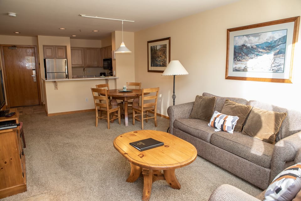 A neatly furnished living room with a grey couch, a wooden coffee table, a dining table with chairs, and a kitchen area in the background. There are framed pictures on the walls.