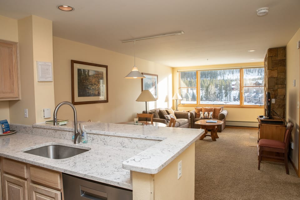 Modern open-plan living space with a kitchen, marble countertops, a living room with sofas, a flat-screen TV, large windows, and a scenic view of a snowy landscape.