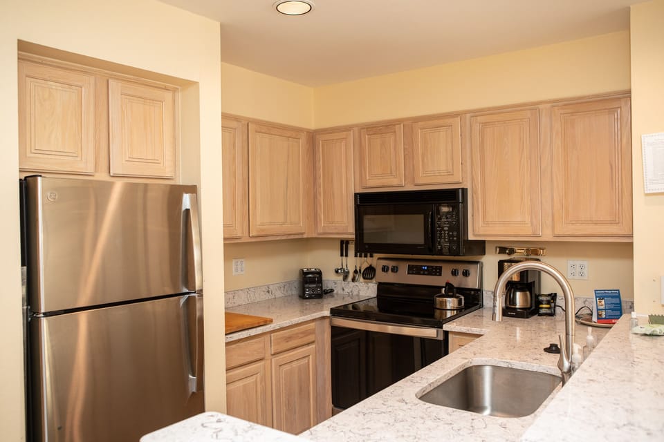 A modern kitchen with light wood cabinets, a black microwave and stove, a stainless steel refrigerator, a coffee maker, and a marble countertop with a sink.