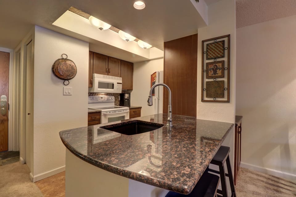 A modern kitchen features granite countertops, a sleek sink with a high-arch faucet, a decorative hanging pot, an electric stove, and a microwave. Three framed artworks adorn the wall.