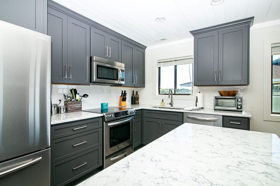 Bright kitchen with gray cabinets, marble-look island, stainless appliances, white backsplash, sink window, toaster oven, and coastal vacation rental style.