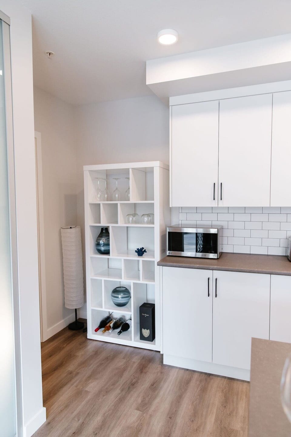 Effortlessly organize in this sleek kitchen with white cabinetry and a stylish shelving unit. Store wine bottles and décor, while the microwave keeps cooking simple. A minimalist design for a fresh feel.