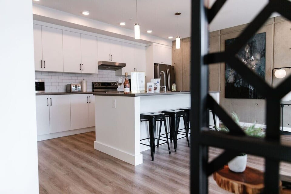 Cook and gather in this sleek kitchen with white cabinets and stainless steel appliances, featuring an island with seating for four. The dining area, framed by a geometric divider, boasts a concrete wall and vibrant art.