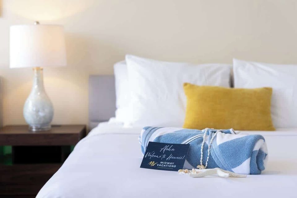 A neatly made bed with a yellow accent pillow and a welcome towel arrangement for a relaxing stay.
