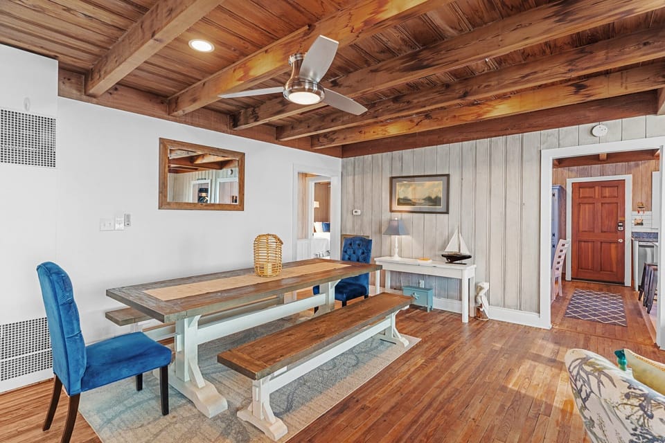 Dining room with a farmhouse table, bench seating, blue chairs, wood floors, cottage wall paneling, and a warm wood beam ceiling.
