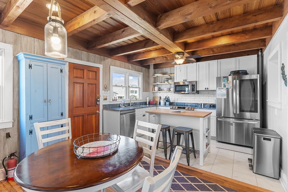 Charming dining space with a round table, white chairs, wood beam ceiling, red accent cabinet, hutch, bookshelves, and beachy cottage decor.