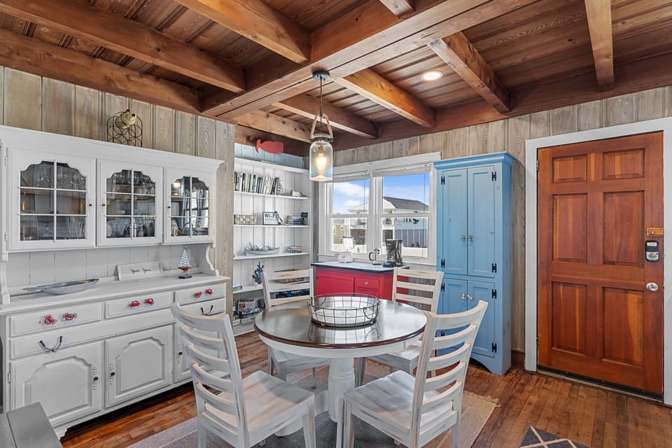 Charming dining space with a round table, white chairs, wood beam ceiling, red accent cabinet, hutch, bookshelves, and beachy cottage decor.