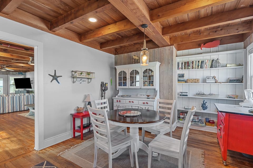 Charming dining space with a round table, white chairs, wood beam ceiling, red accent cabinet, hutch, bookshelves, and beachy cottage decor.