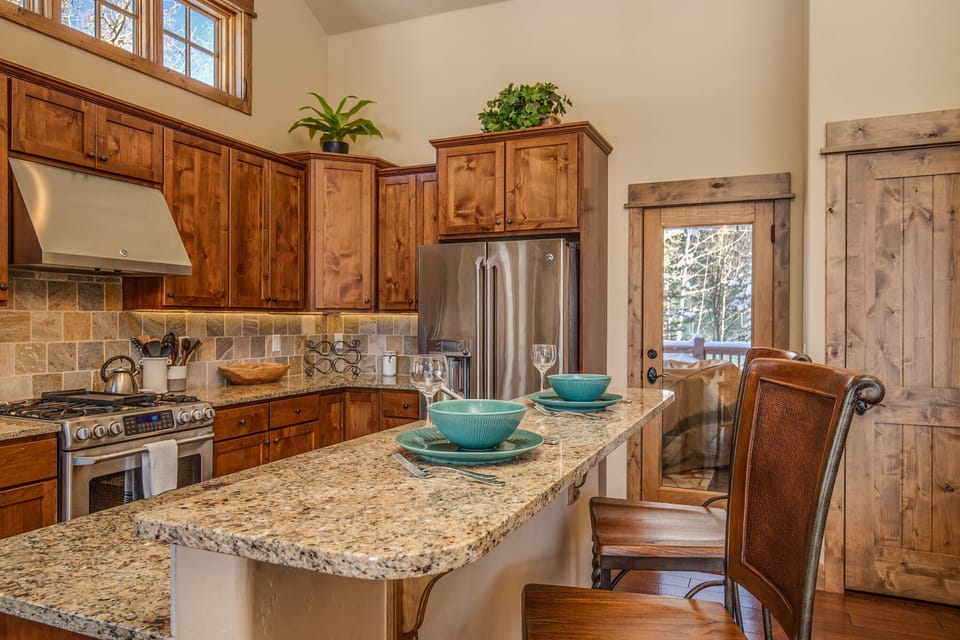 A modern kitchen with wooden cabinets, granite countertops, stainless steel appliances, and a breakfast bar set with blue dinnerware and glasses. There are plants on top of the cabinets and outside light coming through a window and door.