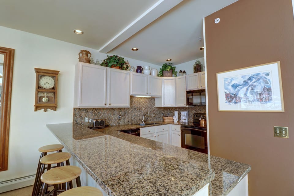 A modern kitchen with granite countertops, white cabinets, and stainless steel appliances. Barstools are lined along the island. Decorative items and plants are placed above the cabinets.