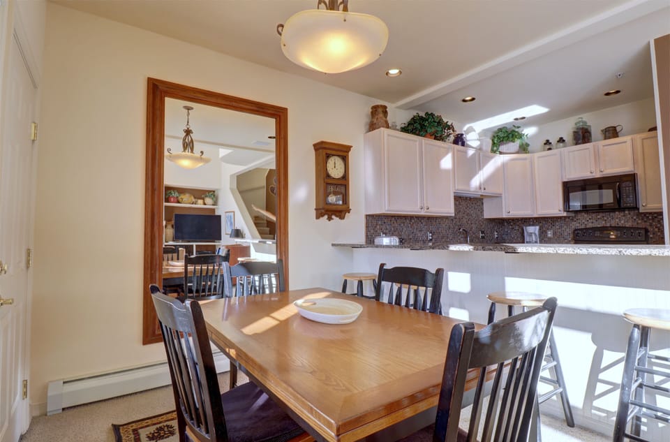 A kitchen and dining area with a wooden table and chairs, a countertop with barstools. Bright lighting from ceiling fixtures illuminates the room.