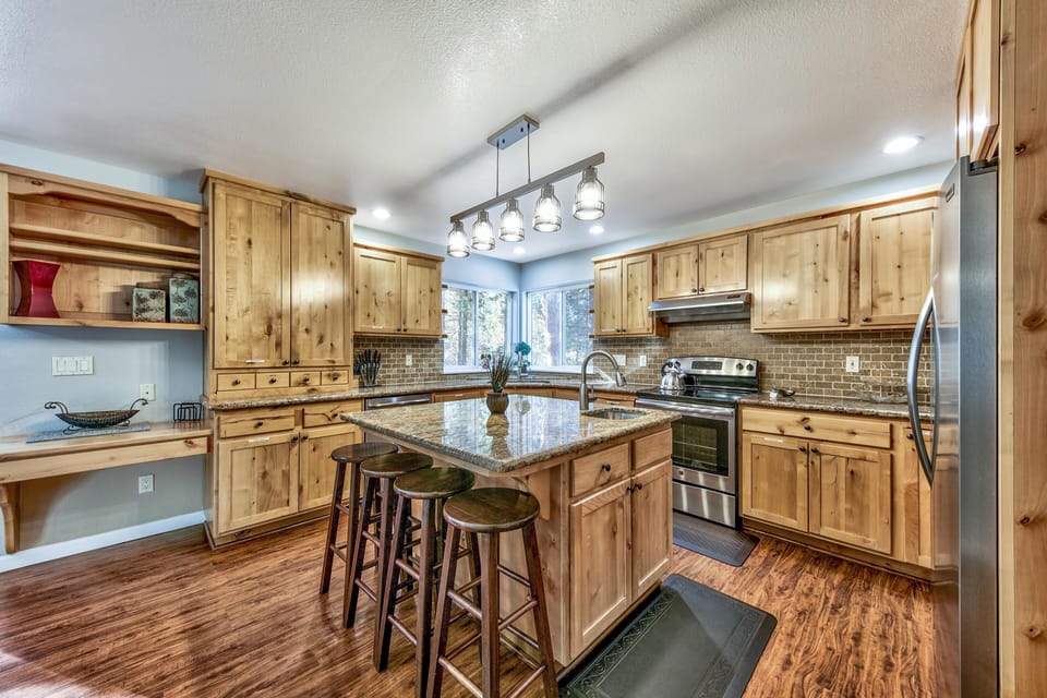 Kitchen features granite island with four wooden stools, knotty pine cabinets, hardwood floors, and corner forest windows.