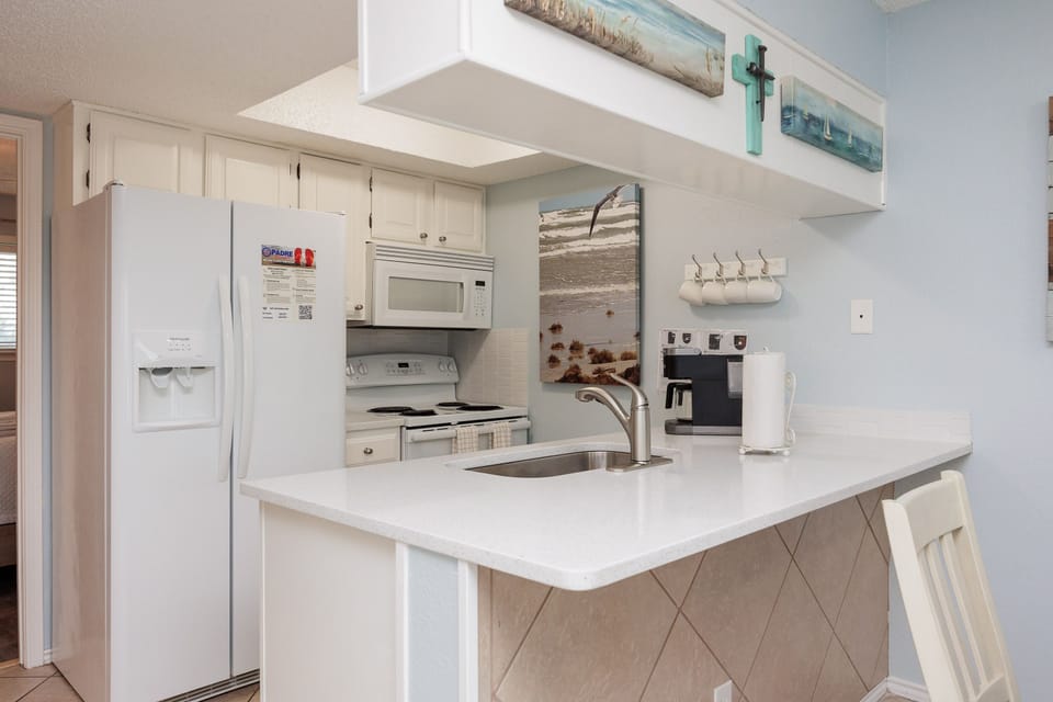 Your fully equipped kitchen features white appliances, quartz countertops, and coastal artwork creating a bright, beach-inspired cooking space.