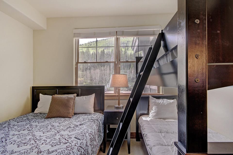 A bedroom featuring a double bed and a bunk bed with a ladder. The room has a large window with shades, offering a view of a forested hillside. A bedside table with a lamp is placed between the beds.