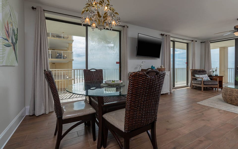 This dining area features a glass table and woven chairs, with a view of the beach and a wall-mounted TV for entertainment