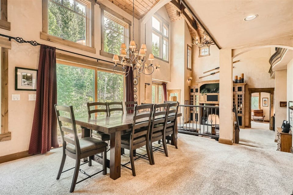 Spacious dining area with a large wooden table, high vaulted ceilings, chandelier, large windows, and carpeted floors, leading to a living room with a balcony and built-in shelves.