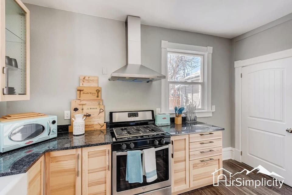 - Bright kitchen featuring a gas range with vent hood, granite counters, and wood cabinetry
- Decorative wine crates add a creative touch above the backsplash and window
- Natural light fills the space for a welcoming and functional cook zone