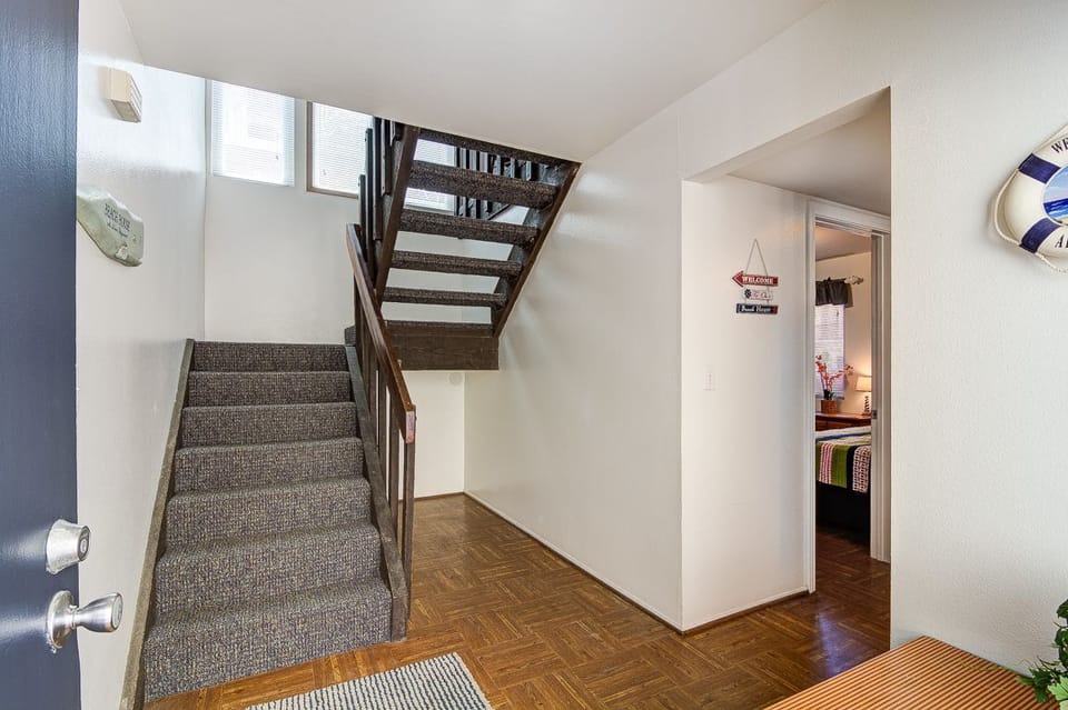 The front entryway of the home, with stairs that lead to the kitchen, living area, and primary bedroom on the left, and a hallway that leads to the remaining bedrooms on the right.
                