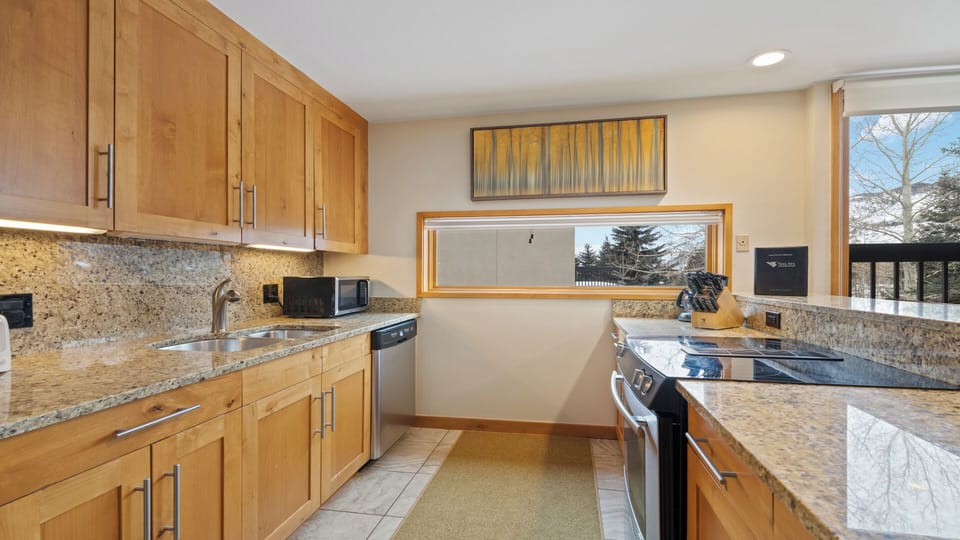 Your fully equipped kitchen features granite countertops, stainless steel appliances, and warm wood cabinetry—perfect for preparing meals while enjoying natural light and tree views through large windows.