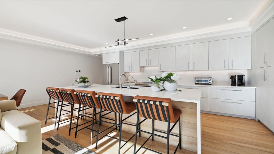 Gather around the spacious kitchen island where sunlight streams across sleek white cabinetry and warm wood finishes, creating the perfect spot for morning coffee or evening conversation with loved ones.