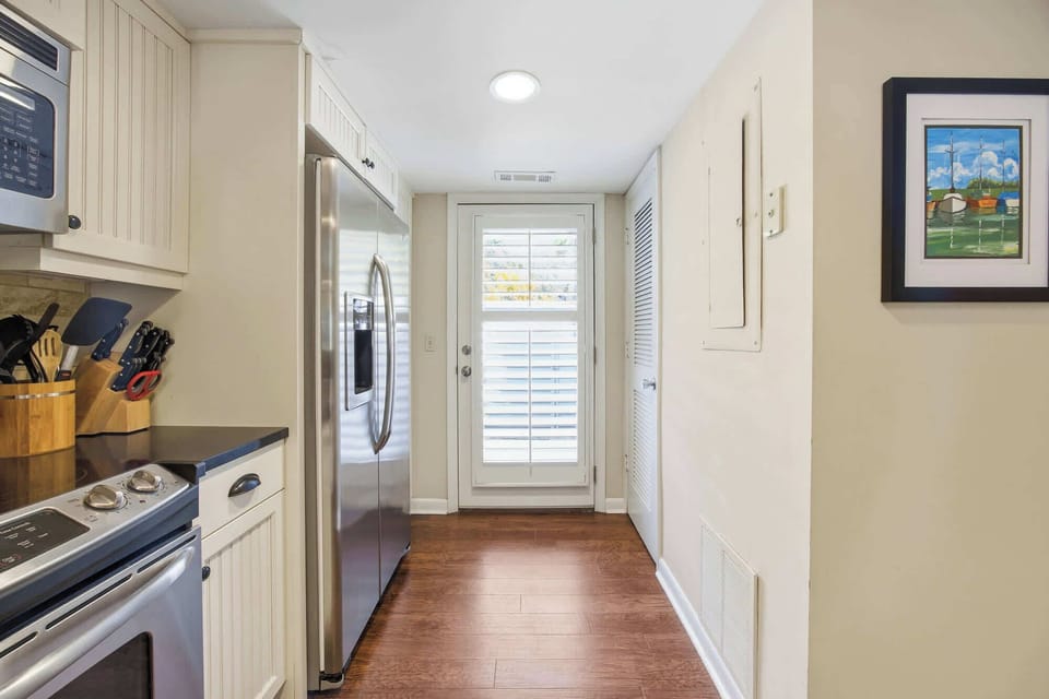 Stainless steel appliances gleam in this efficient kitchen, where rich wood floors lead your eye toward a sun-washed door. Shutter-filtered rays brighten the clean white cabinetry and nautical art, creating an airy spot for morning coffee.