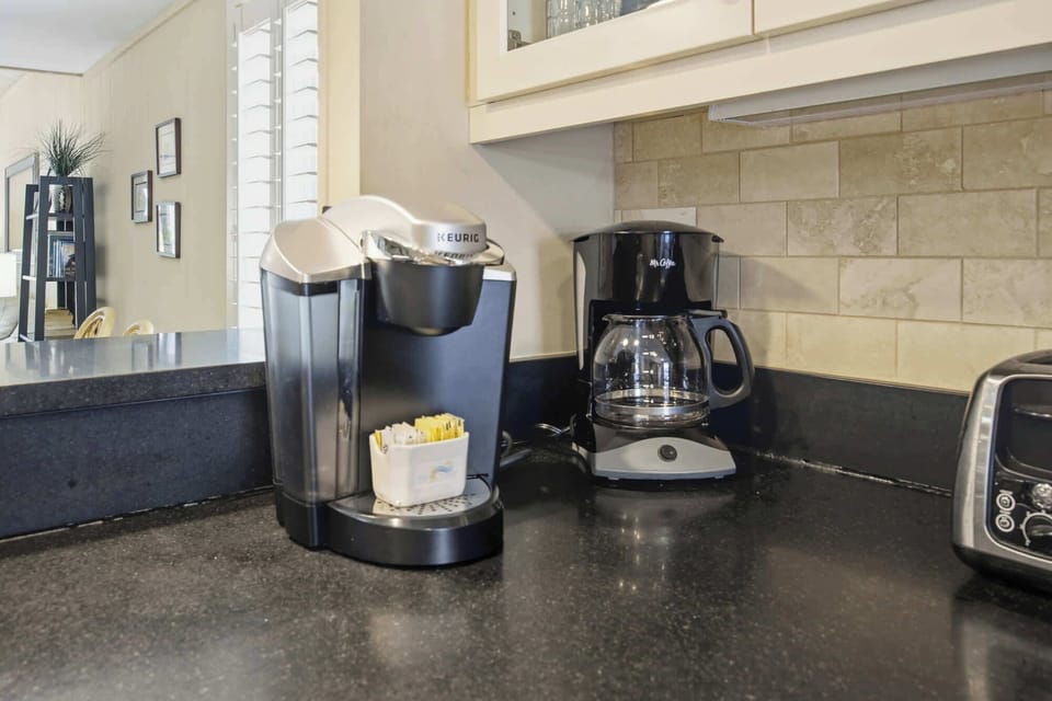 Dark counters anchor this convenient coffee corner, where earthy tiles and bright cabinetry create a grounded, functional space. Take your pick between a quick brew or a full pot to start your morning in this calm, well-equipped kitchen.