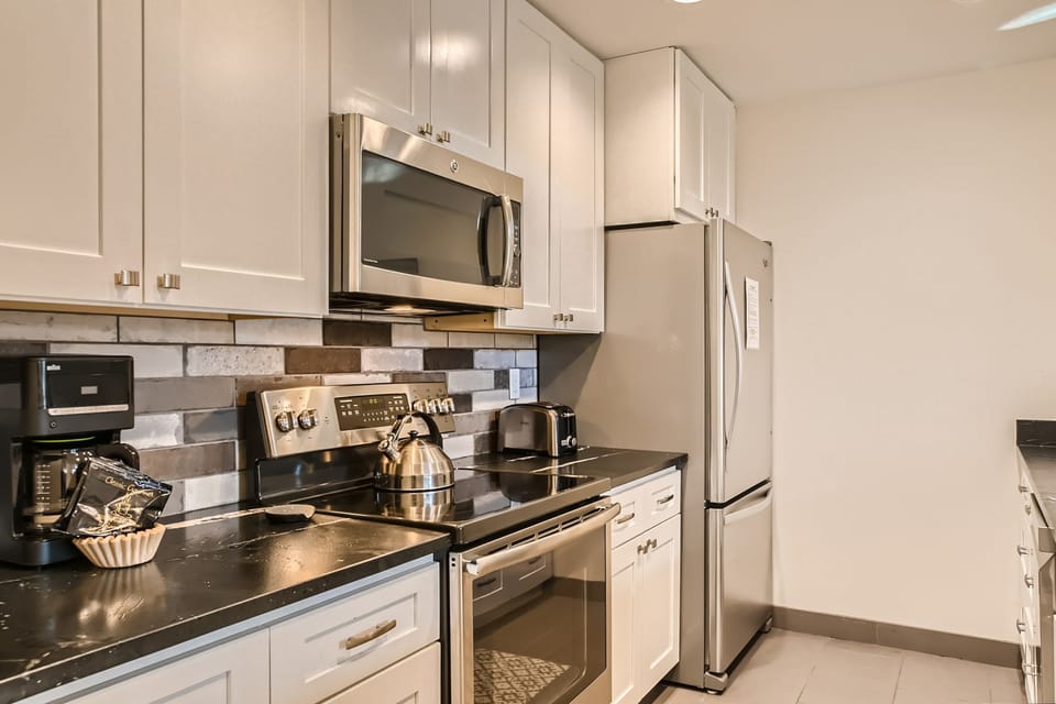 Modern kitchen with stainless steel appliances, including a microwave, stove, and refrigerator. White cabinets, black countertops, and a coffee maker are also present.