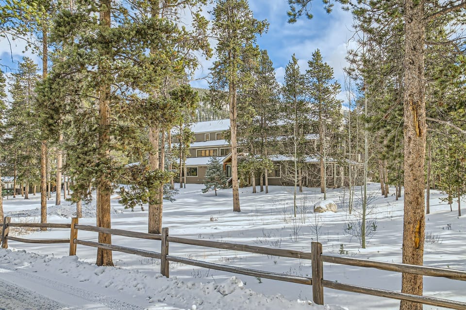 A building surrounded by snow-covered trees and ground, with a wooden fence in the foreground on a clear, sunny day.