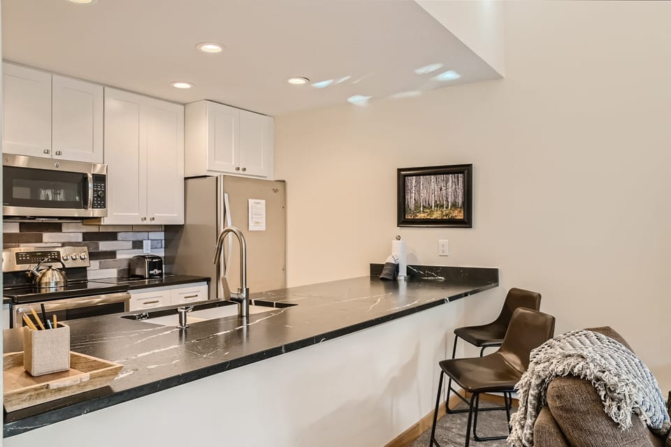 Modern kitchen with black marble countertop, stainless steel appliances, and white cabinetry. There are two barstools with a blanket on one, and a framed picture on the wall.