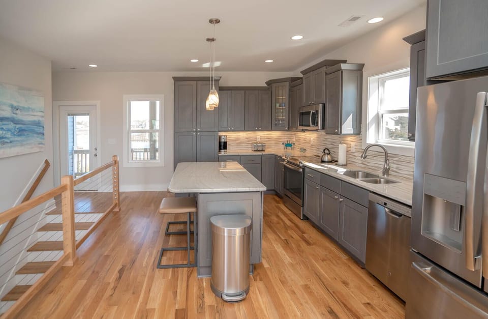 Bright kitchen with gray cabinets, stainless appliances, island seating, pendant lights, wood floors, large windows, and marsh-view style coastal decor.