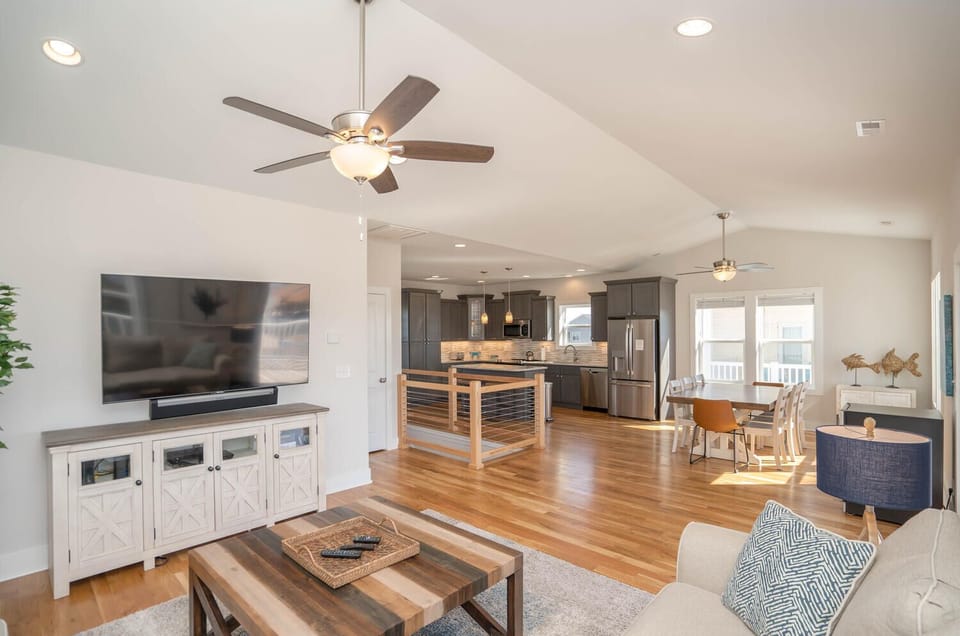 Open living room with cream sofas, wood floors, vaulted ceiling, ceiling fan, TV, coffee table, coastal decor, large windows, and balcony access.