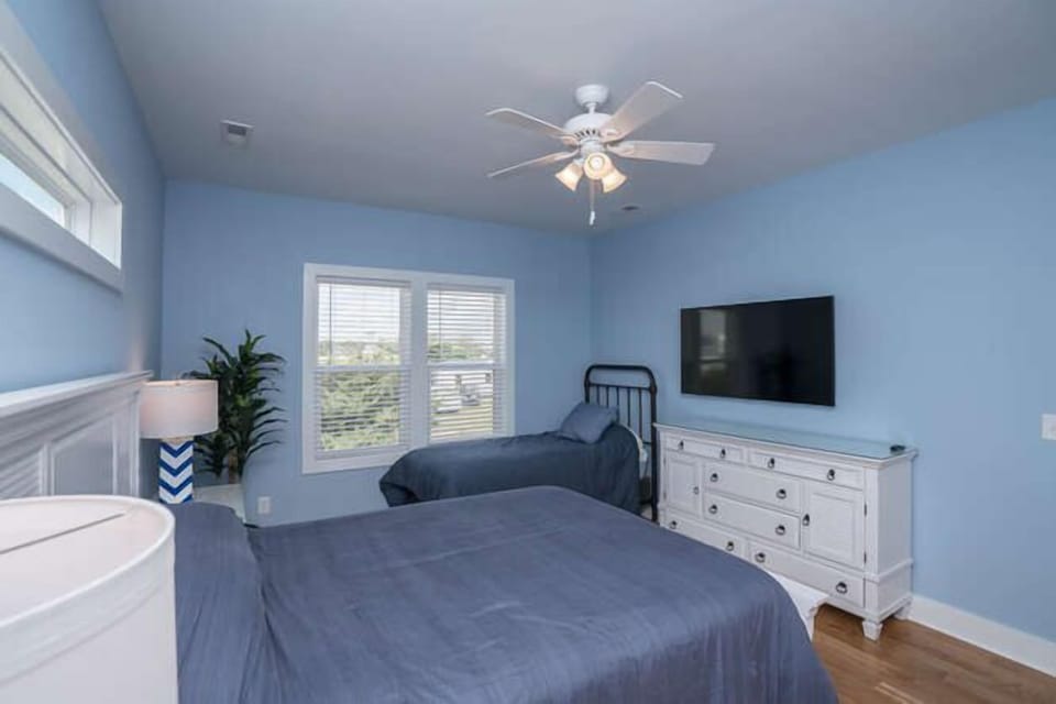 Bedroom with blue walls, two beds, wall-mounted TV, white dresser, ceiling fan, window blinds, bedside lamp, plant, wood floors, and coastal rental style.