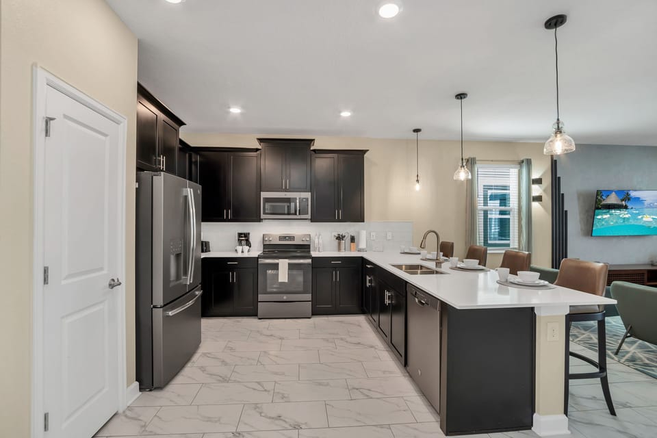 A sleek, modern kitchen with dark cabinetry and a bright, spacious layout - Stainless steel appliances and a large island make it a dream for any chef - This kitchen’s open design is perfect for cooking and connecting with loved ones