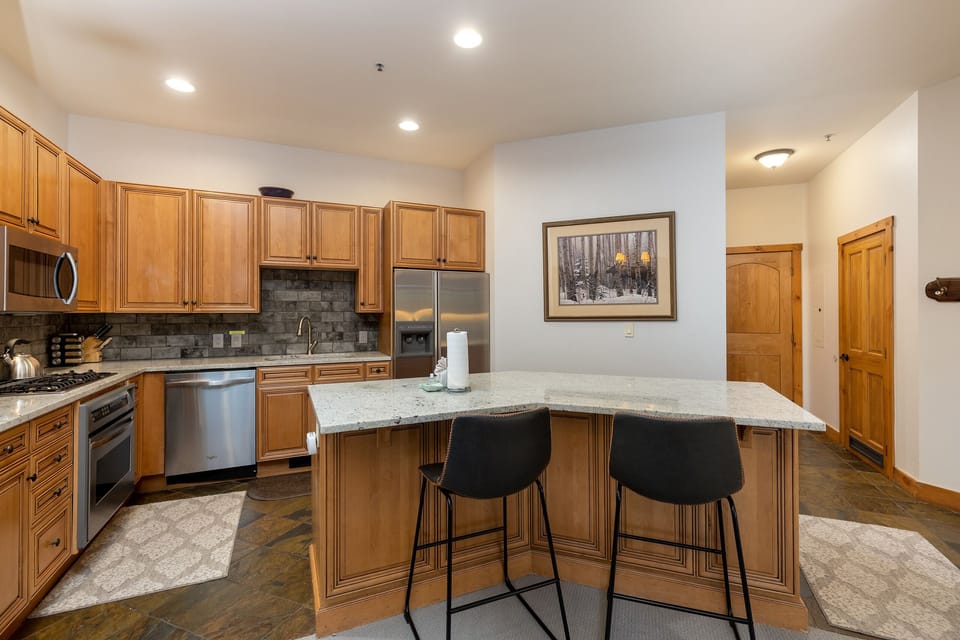 Modern kitchen with wooden cabinets, stainless steel appliances, granite countertops, and a central island with two black barstools. There is a framed landscape picture on the wall.