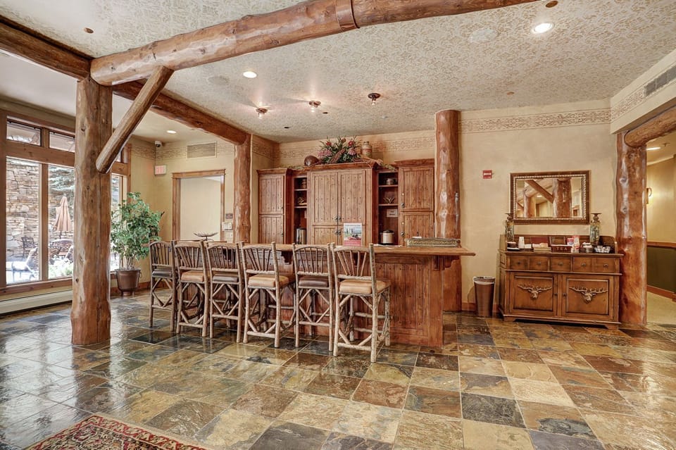 A rustic reception area features a wooden check-in counter with high chairs, a decorative cabinet, and a mirror. The space has stone tile flooring and large windows providing natural light.