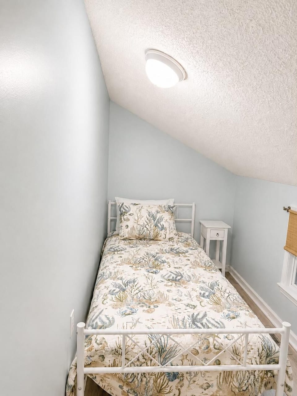 Single white metal bed with ocean-themed bedding fits under sloped ceiling beside a small white nightstand.