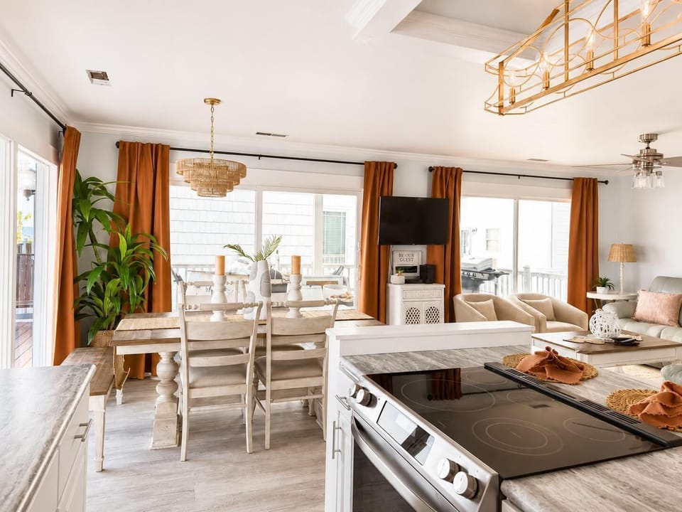 Whitewashed wood dining table with mixed seating, wicker chandelier, and sliding glass doors with rust curtains.