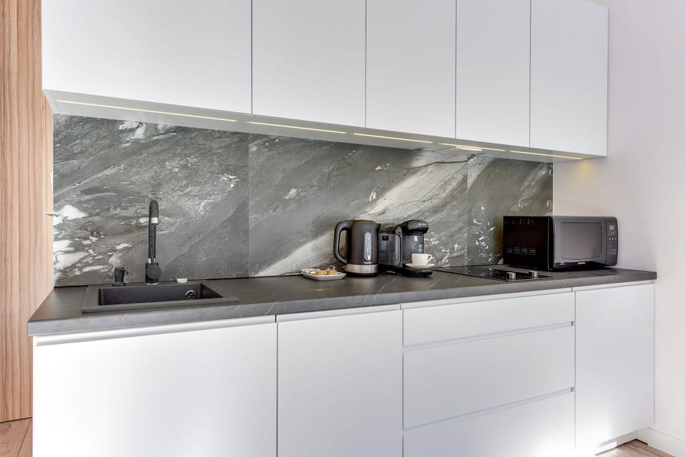 A functional kitchenette with white cabinets, a marble countertop, and a black backsplash. Large windows brighten the space.