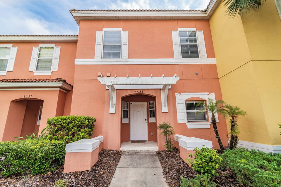 Step into a charming exterior framed by lush landscaping, where the warm peach tones of the home invite you to unwind. Large windows allow natural light to flood the entrance, creating a welcoming atmosphere for your stay.